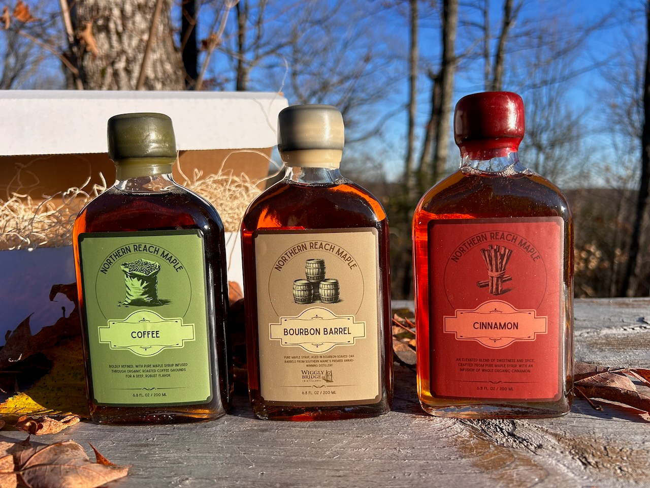 Bottles of Northern Reach Maple's coffee infused, cinnamon infused, and bourbon barrel aged maple syrups with fall leaves in the background.