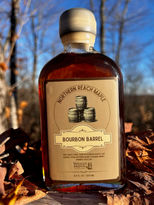 A bottle of Northern Reach Maple's bourbon barrel aged maple syrup with fall leaves in the background.