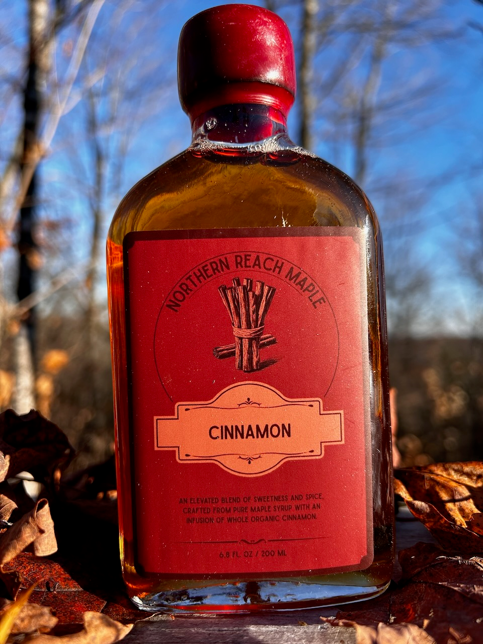 A bottle of Northern Reach Maple's cinnamon infused maple syrup with fall leaves in the background.