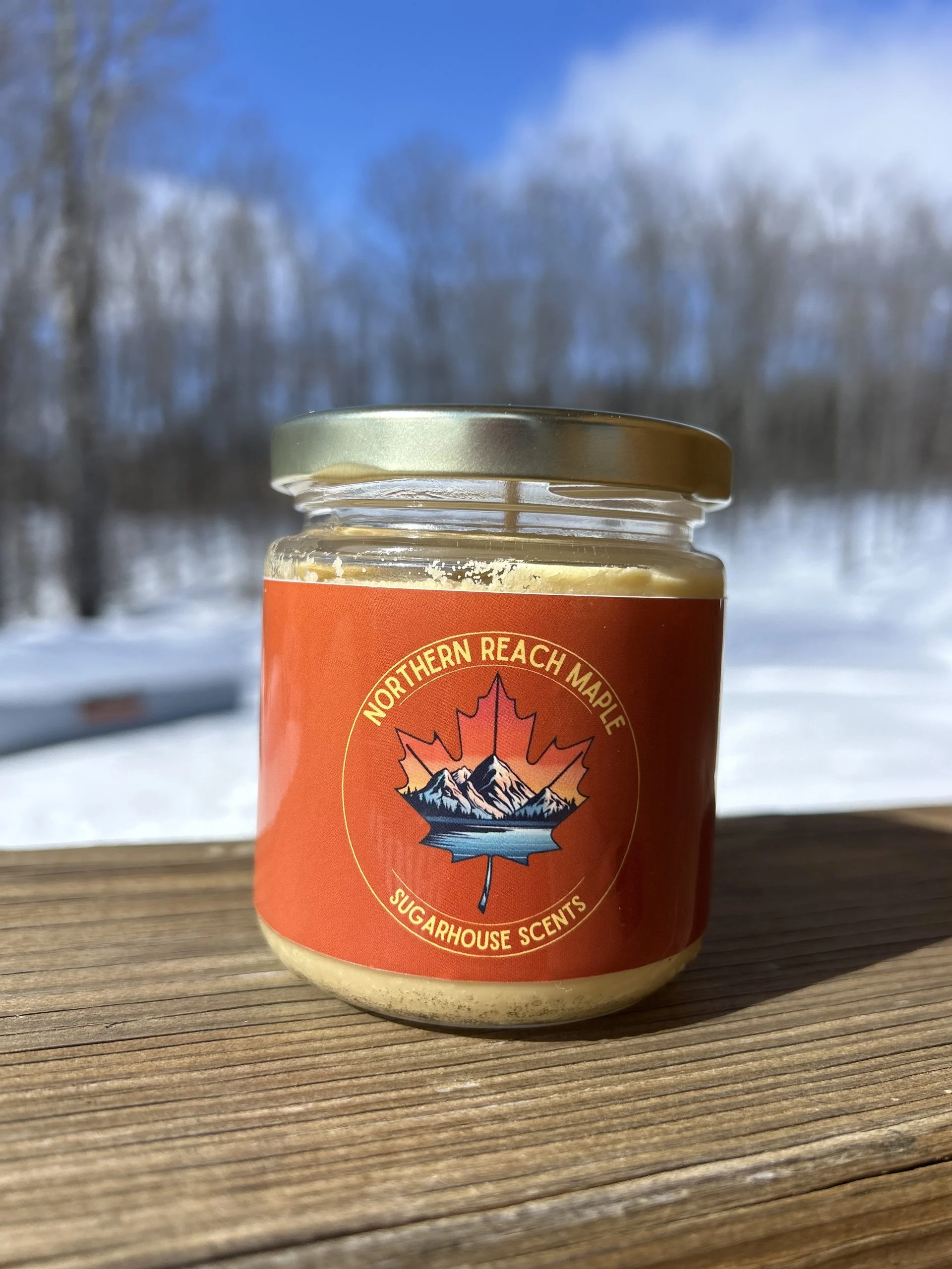 A picture of a maple scented candle with bare trees and snow in the background.