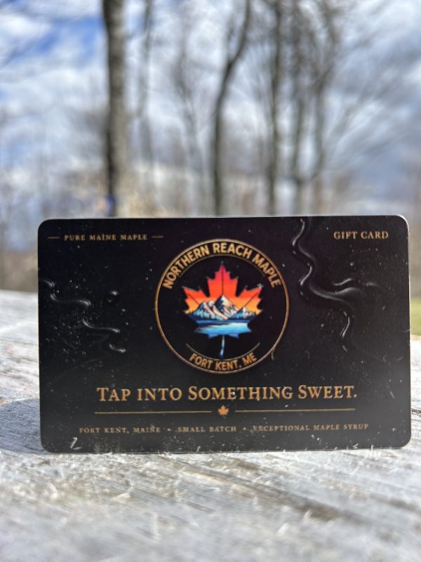 A black and gold Northern Reach Maple Gift Card on a piece of distressed lumber with maples and a blue and cloudy sky in the background.