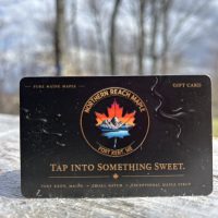 A black and gold Northern Reach Maple Gift Card on a piece of distressed lumber with maples and a blue and cloudy sky in the background.