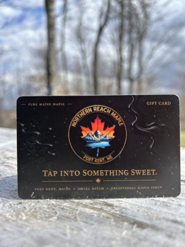A black and gold Northern Reach Maple Gift Card on a piece of distressed lumber with maples and a blue and cloudy sky in the background.