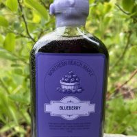 A bottle of blueberry infused maple syrup on a rock in front of a blueberry bush.