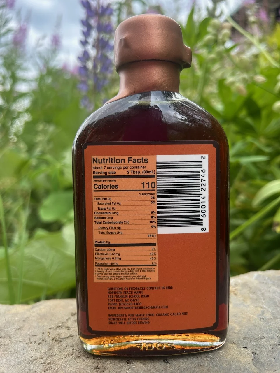 The backside (including the nutrition facts panel) of Northern Reach Maple Cocoa Infused Maple Syrup pictured in front of some lupins in Northern Maine.